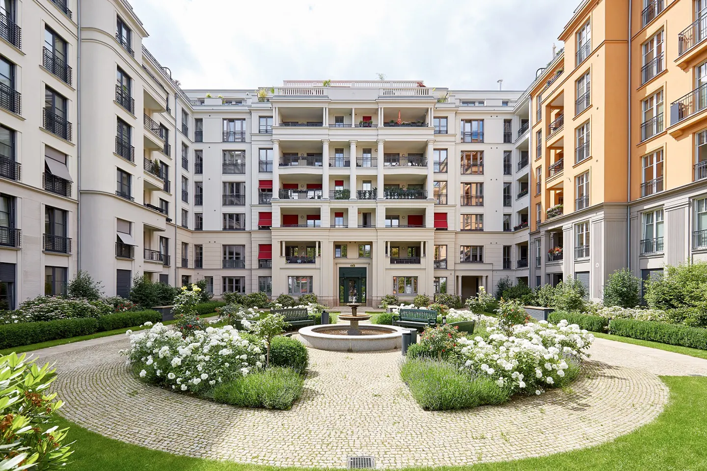 Courtyard view of a multi-story apartment complex with a central fountain and garden. Buildings are white and orange with balconies and windows.