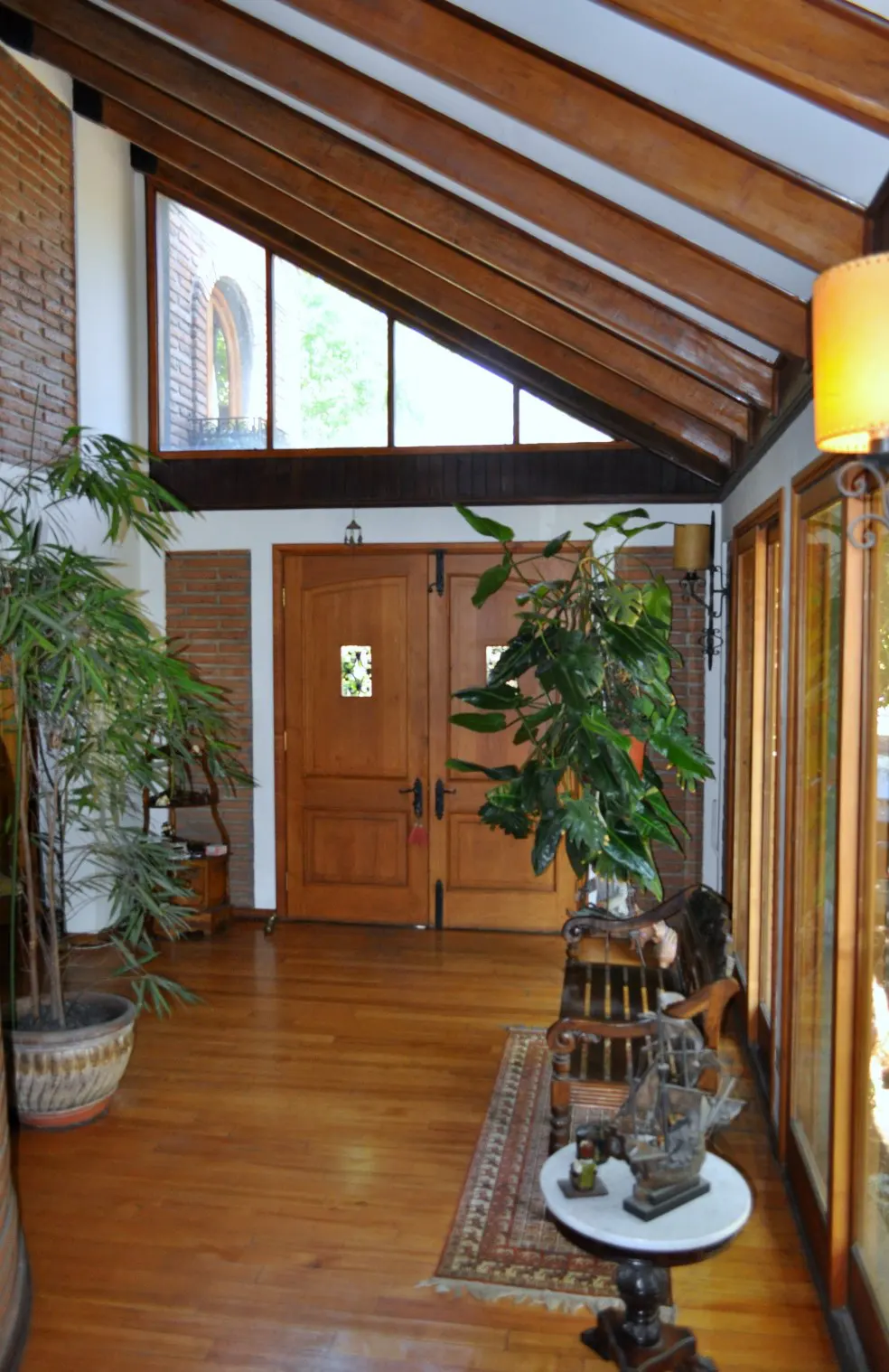 A bright foyer with wood floors, exposed beams, and large windows. Potted plants and a decorative table add to the inviting atmosphere.