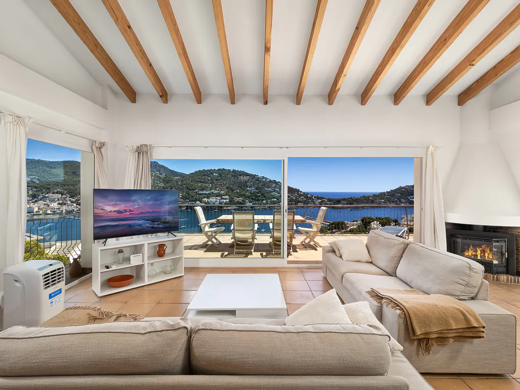 Bright living room with beige sofas, white walls, and wood-beamed ceiling. Large windows offer a scenic view of the ocean and mountains.