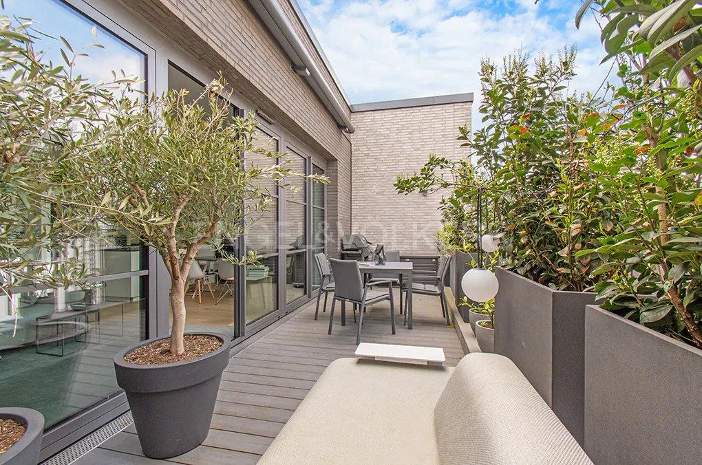 A modern rooftop terrace with gray wood flooring, a dining set, and lush greenery in planters. A beige sofa is in the foreground.