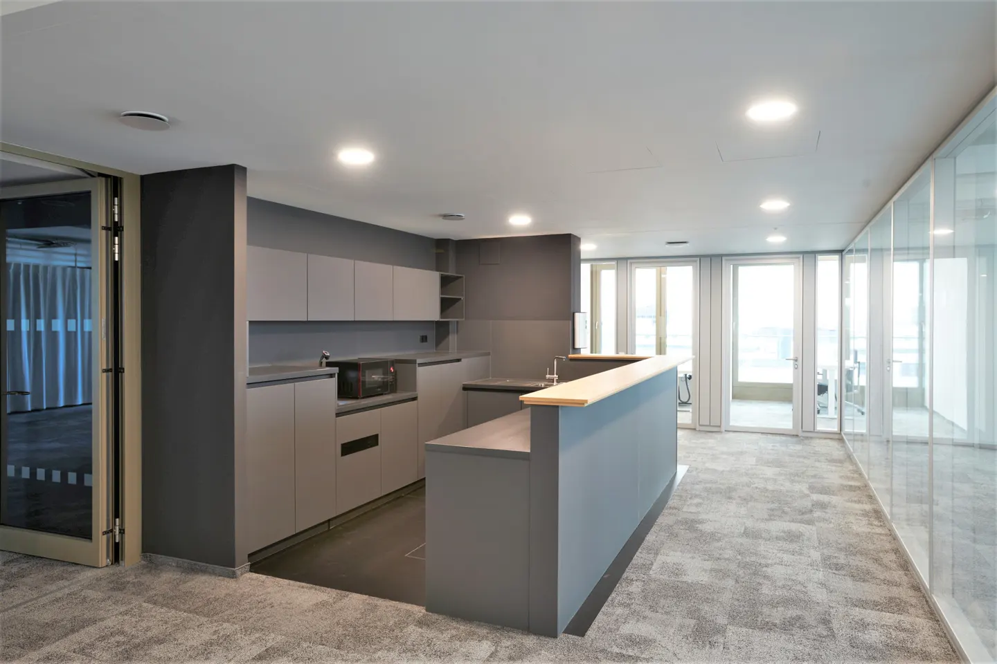 Office break room with gray cabinets, a microwave, and a wooden-topped counter. Glass-walled offices are visible in the background.