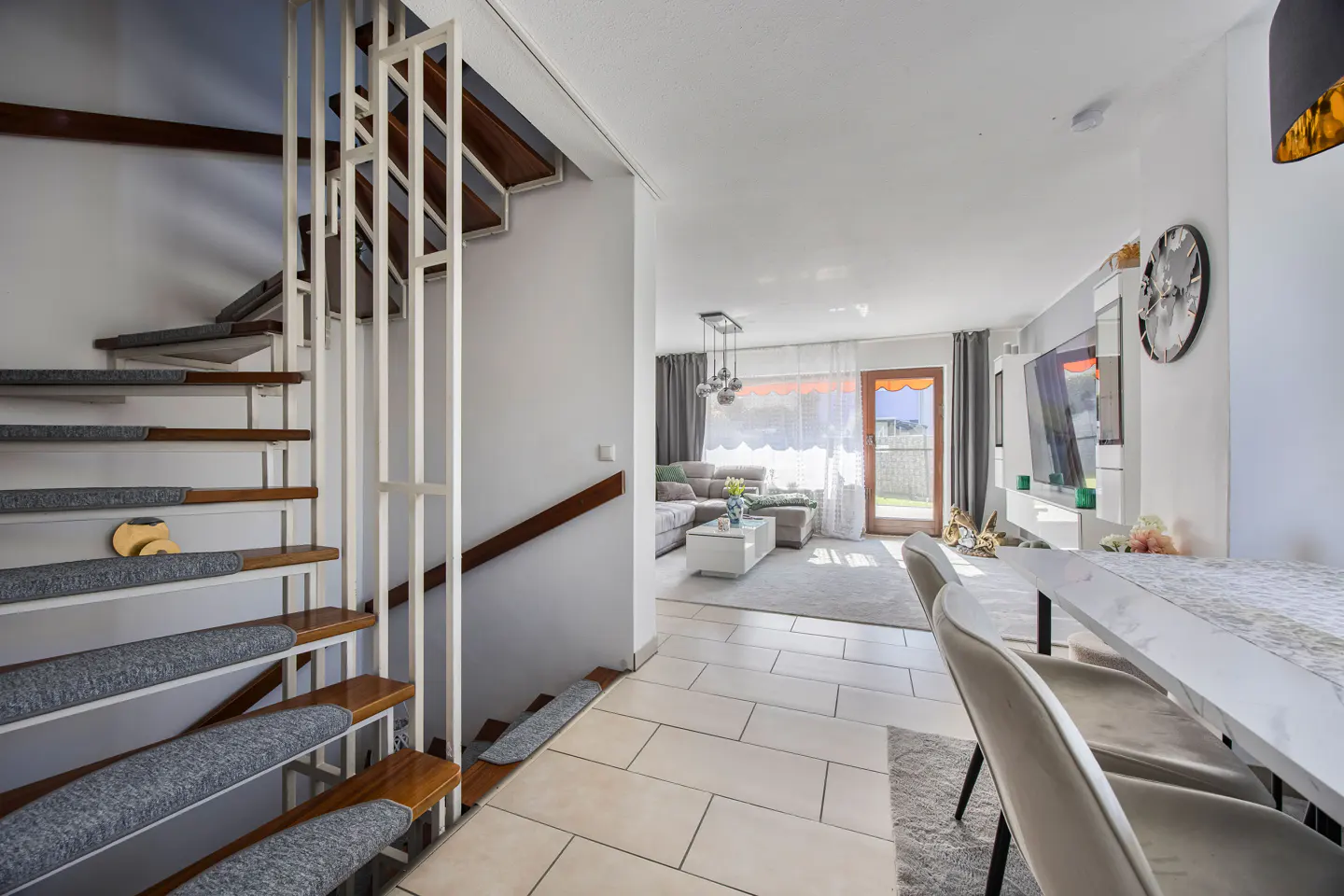 Interior view of a modern home with a staircase, living room, and dining area. The stairs have wooden steps with gray carpet and a white railing.