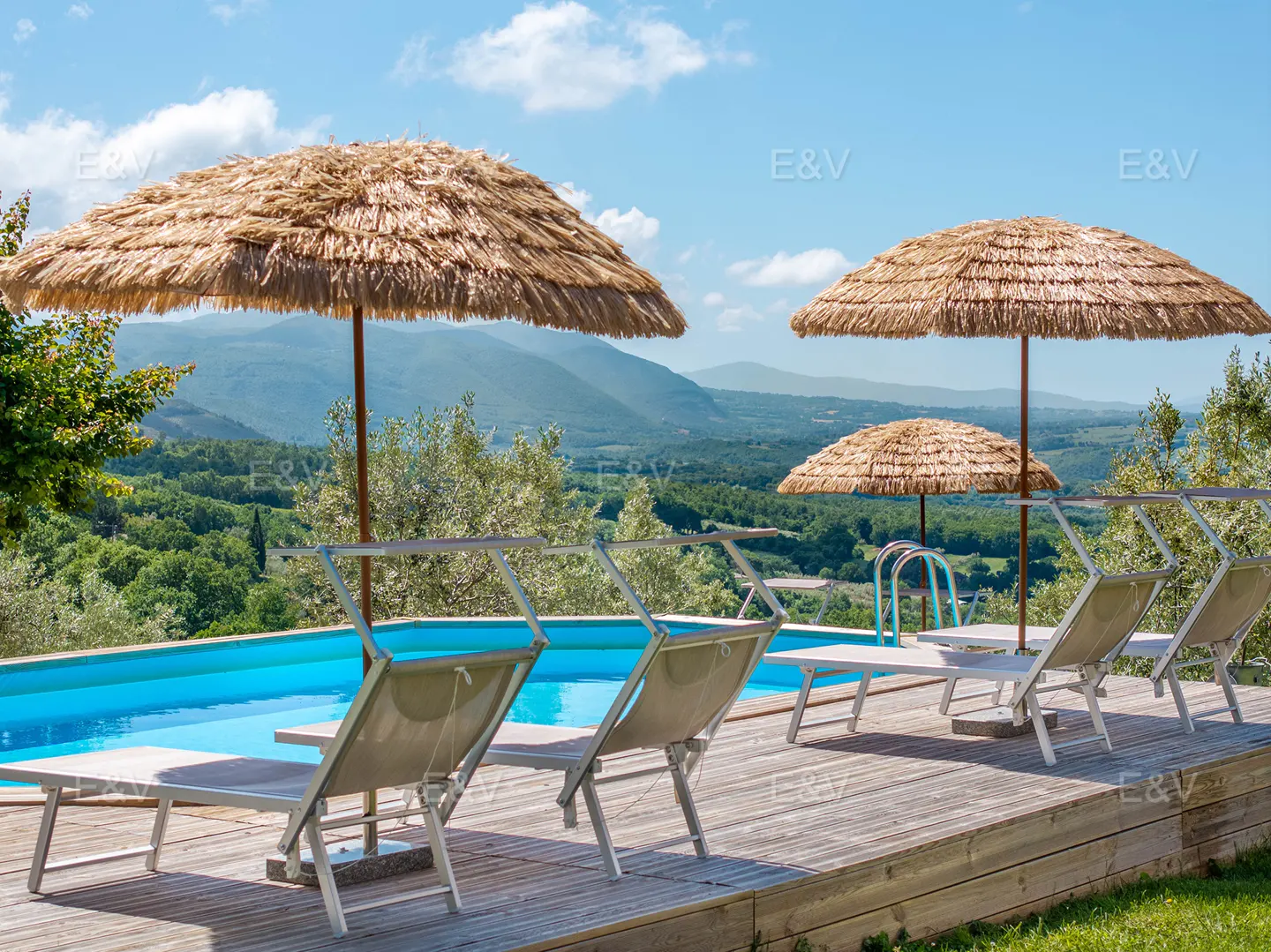 Swimming pool with wooden deck, lounge chairs, and straw umbrellas overlooking a scenic green valley and distant mountains.