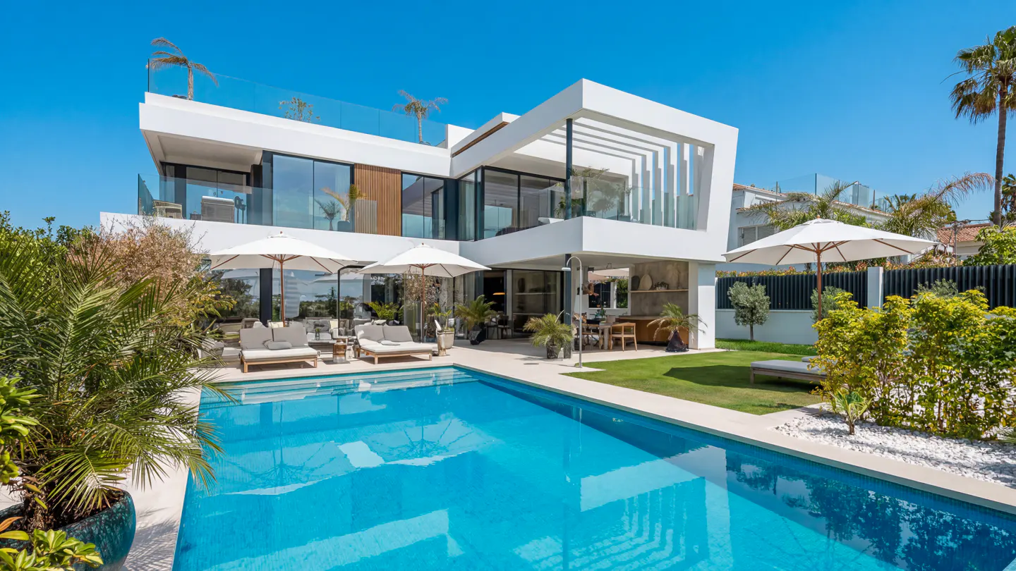 Modern white villa with a blue pool, lounge chairs, and umbrellas under a clear blue sky.