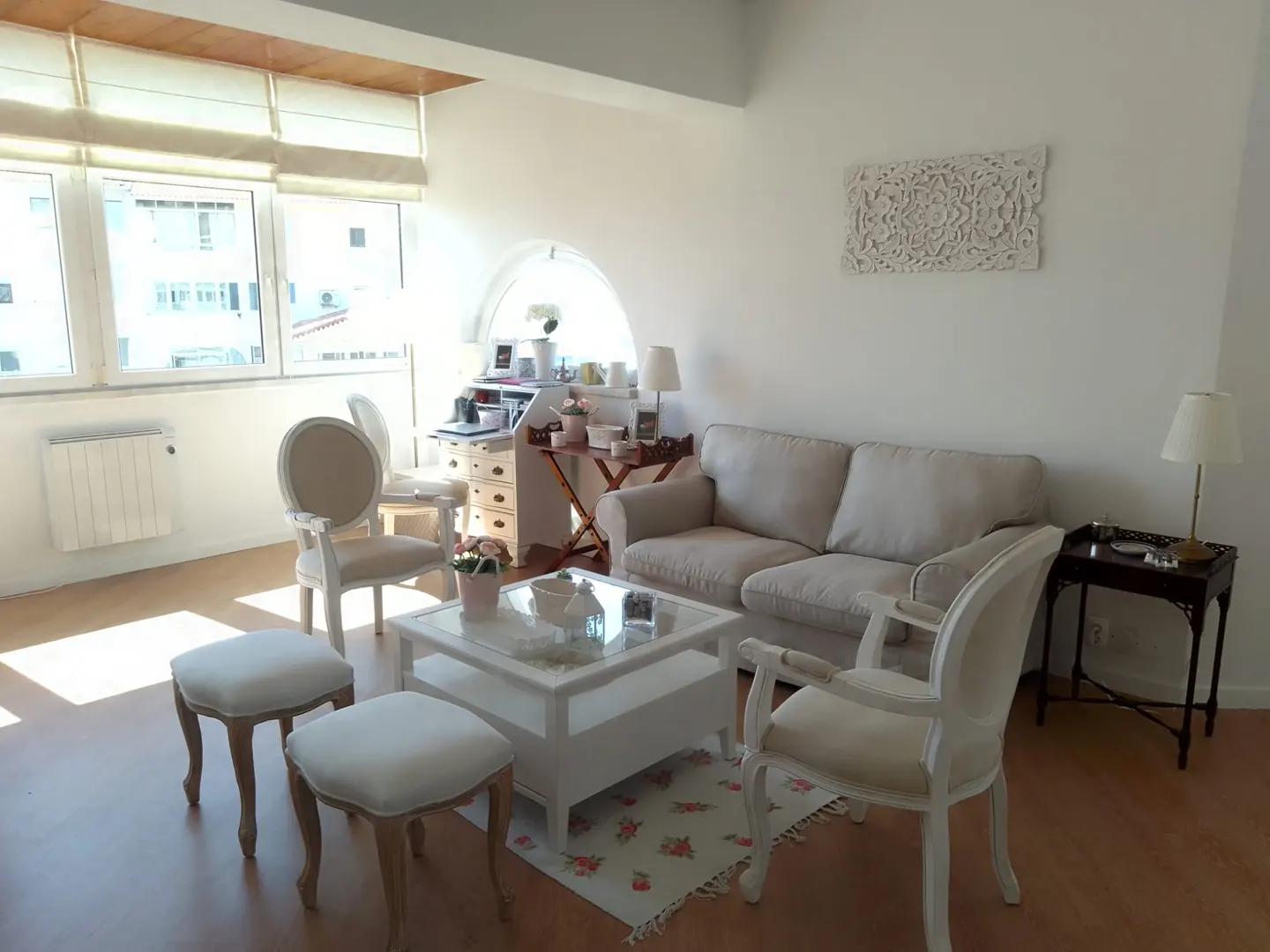 Bright living room with white walls, a beige sofa, white chairs, and a white coffee table on a floral rug. A desk sits by a window.