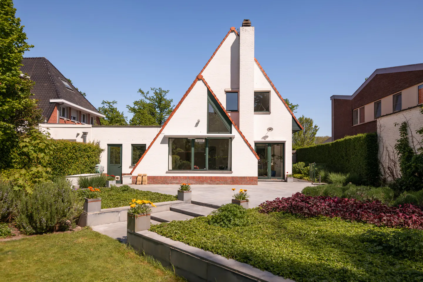 A modern white house with a triangular roof and chimney, surrounded by a lush green garden with flowers and stone steps.