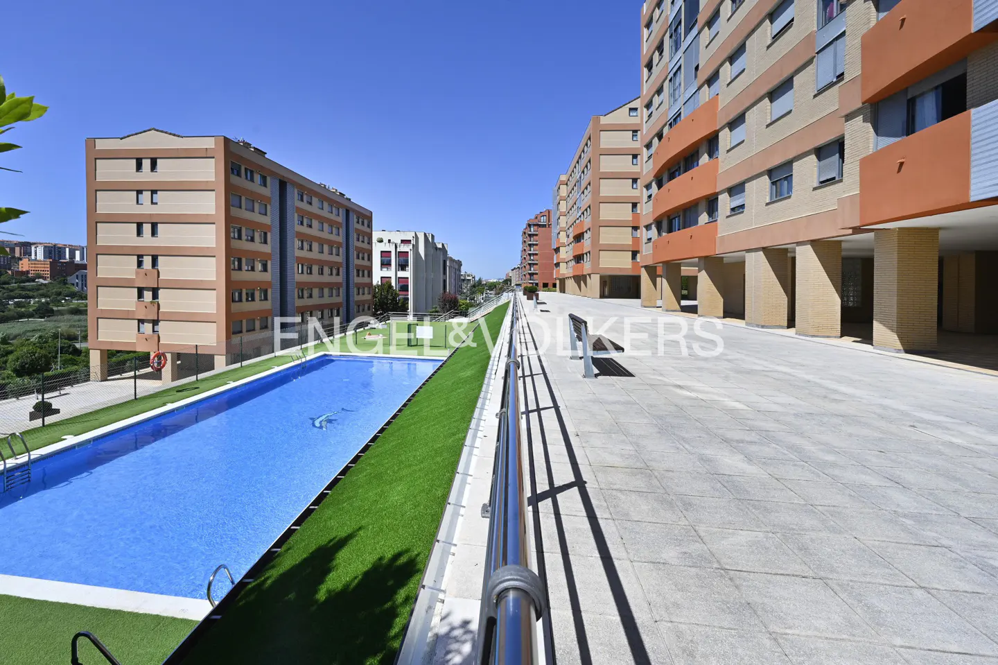 Apartment complex with a blue swimming pool, green turf, and a gray concrete walkway under a clear blue sky.
