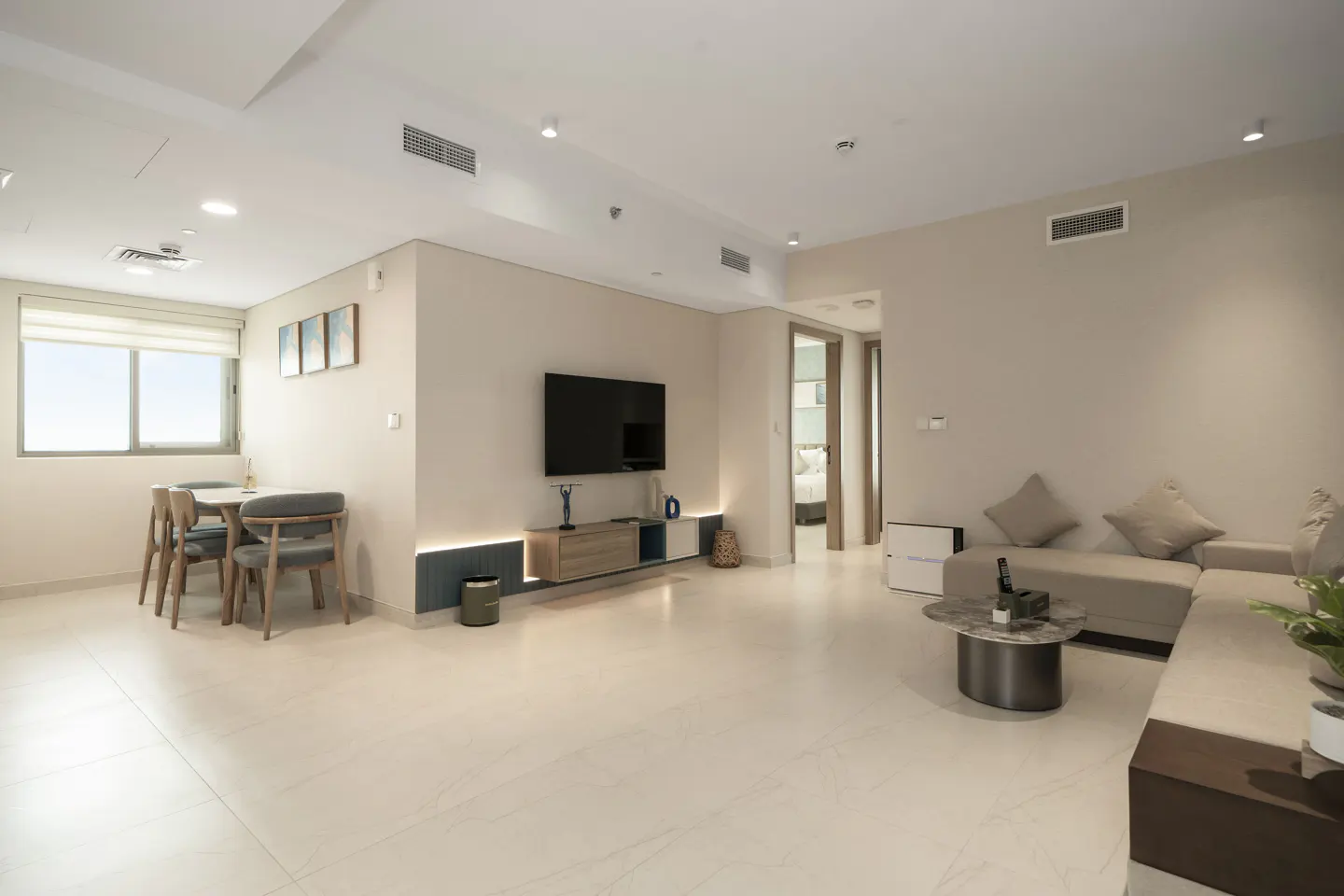 Bright, modern living room with beige walls, tile floors, and a dining table near a window. A TV and sectional sofa furnish the space.