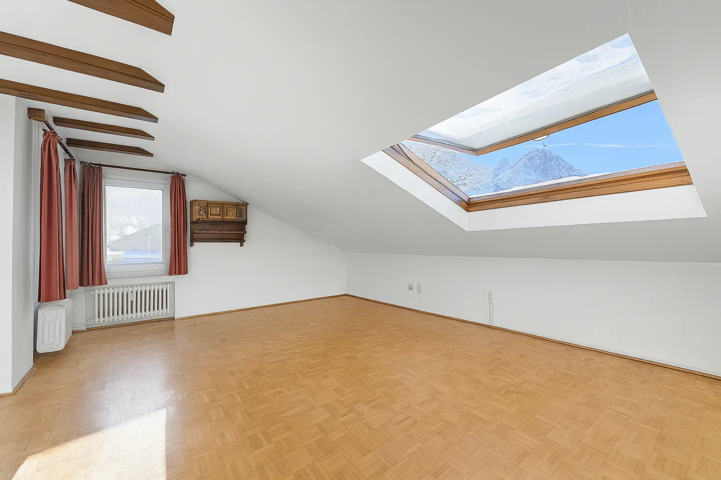 Attic room with wood floors, white walls, and exposed beams. A skylight reveals a mountain view. Curtains frame a window with a radiator.
