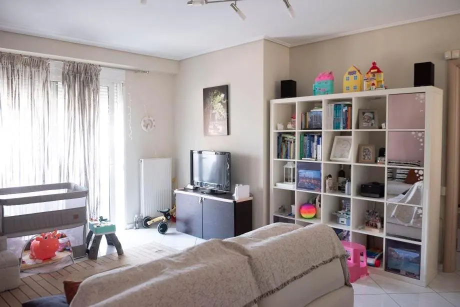 Bright living room with a crib, TV, and white shelving unit filled with books and toys. Beige walls and curtains add warmth.