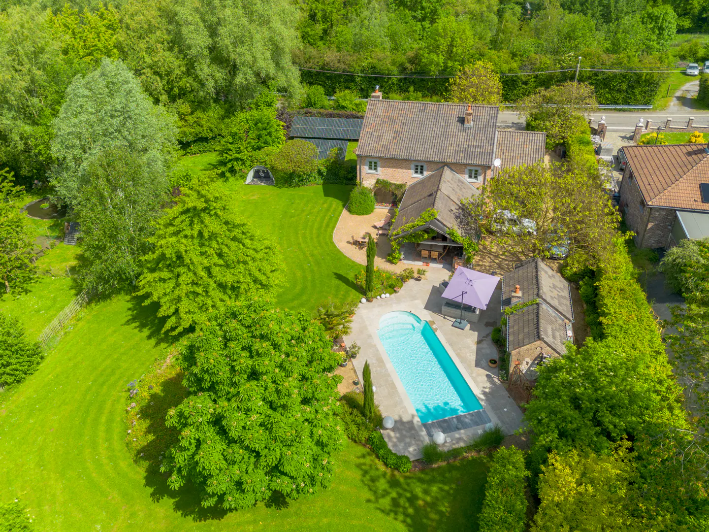 Aerial view of a brick house with a pool, green lawn, and trees. A purple umbrella is near the pool.