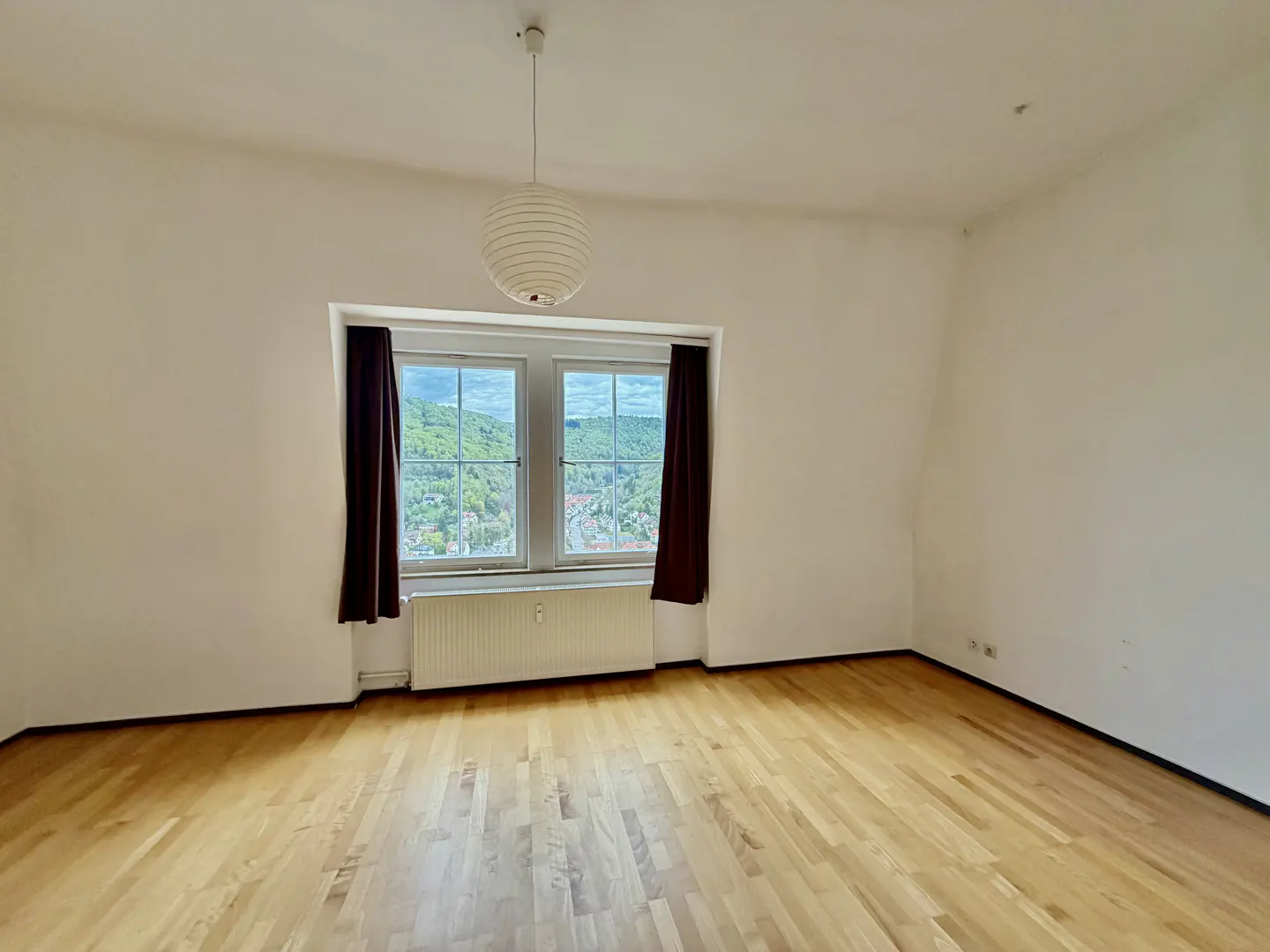 Empty room with wood floor, white walls, and a window with brown curtains. A white radiator is under the window, and a globe light hangs from the ceiling.