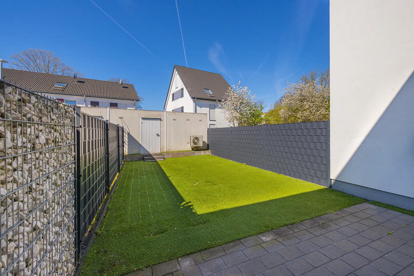 A small backyard with artificial grass, surrounded by fences and a stone wall, under a clear blue sky.