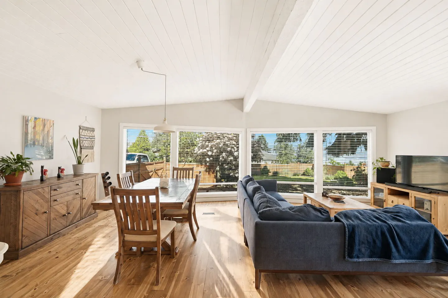 Open-concept living room with hardwood floors, a dark blue sofa, wooden dining set, and large windows with garden views.