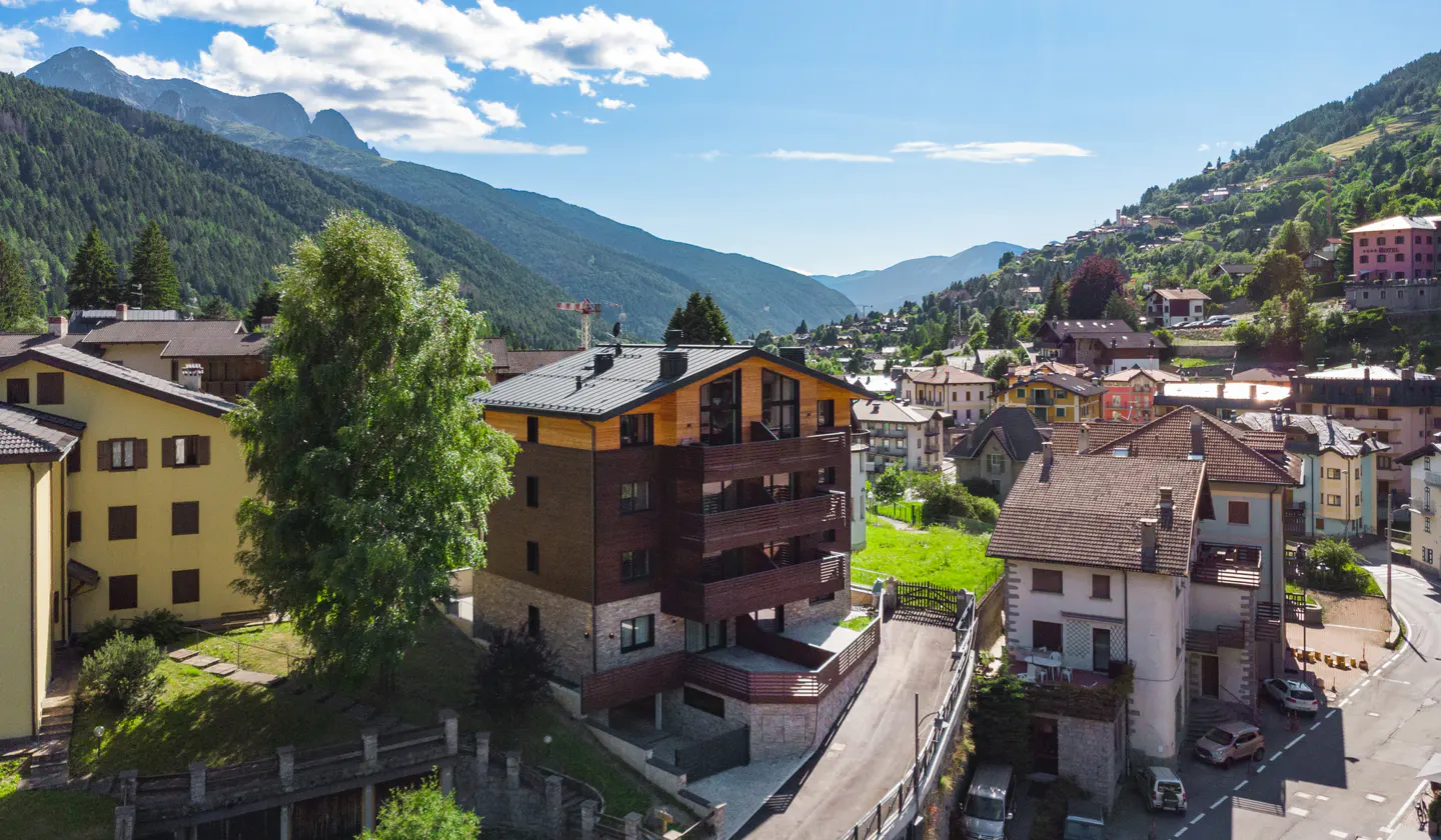 Aerial view of a modern brown and tan building with balconies, nestled in a village surrounded by green mountains under a blue sky.