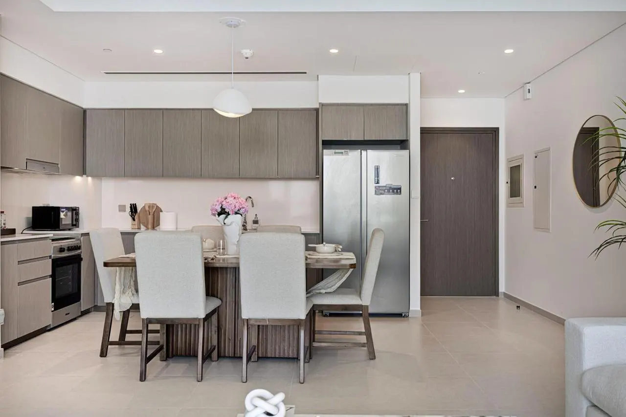 A modern apartment features a dining table with white chairs, a kitchen with gray cabinets, and a stainless steel refrigerator.