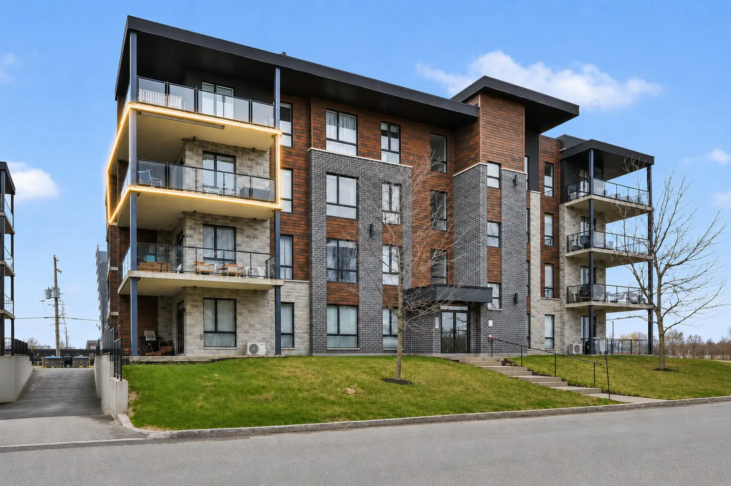 Exterior view of a modern apartment building with brick, stone, and wood facade, balconies, and green lawn.