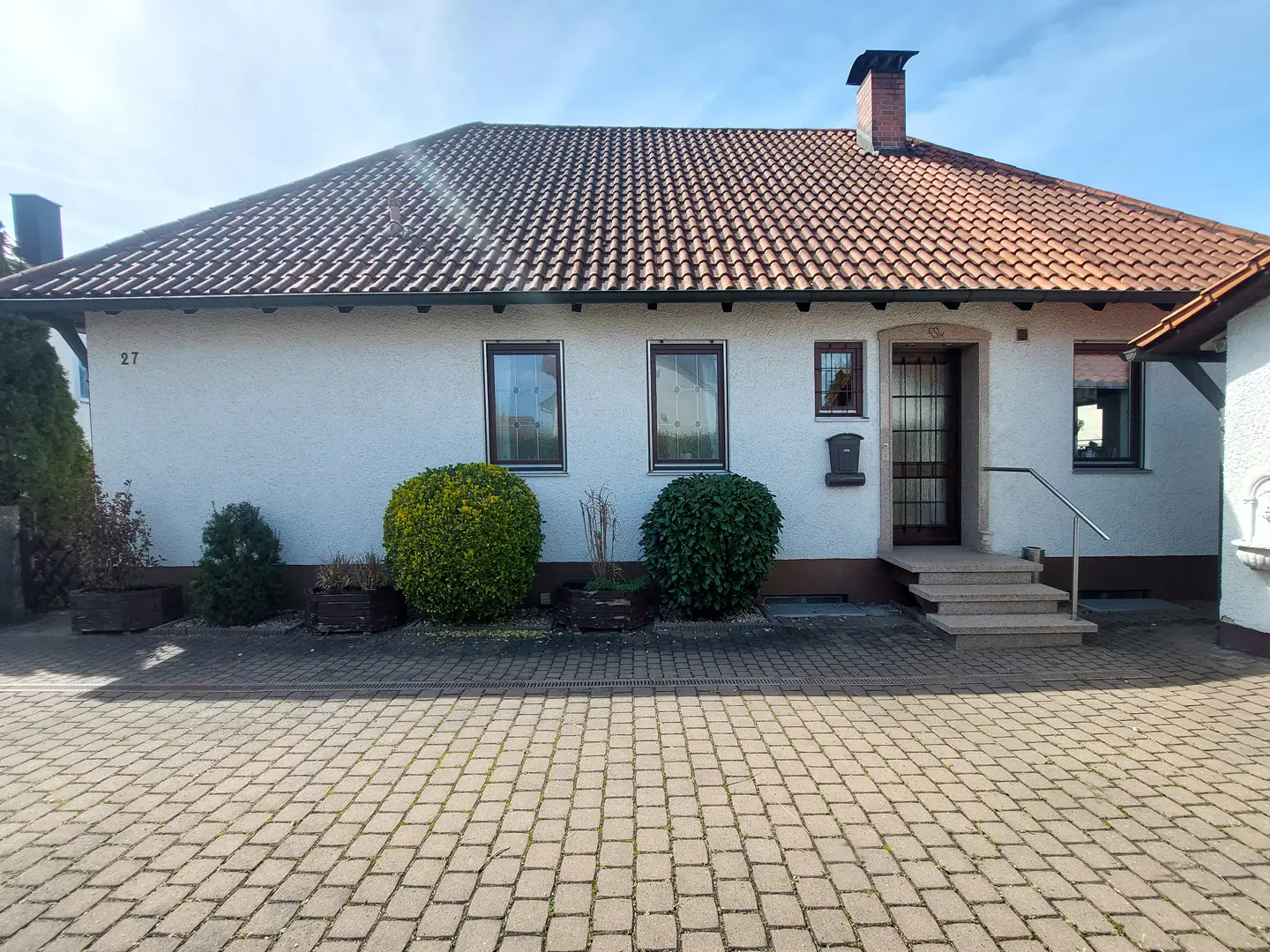 A single-story white house with a red tile roof, brown trim, and a brick driveway. The house has a chimney, several windows, and a front door with steps.