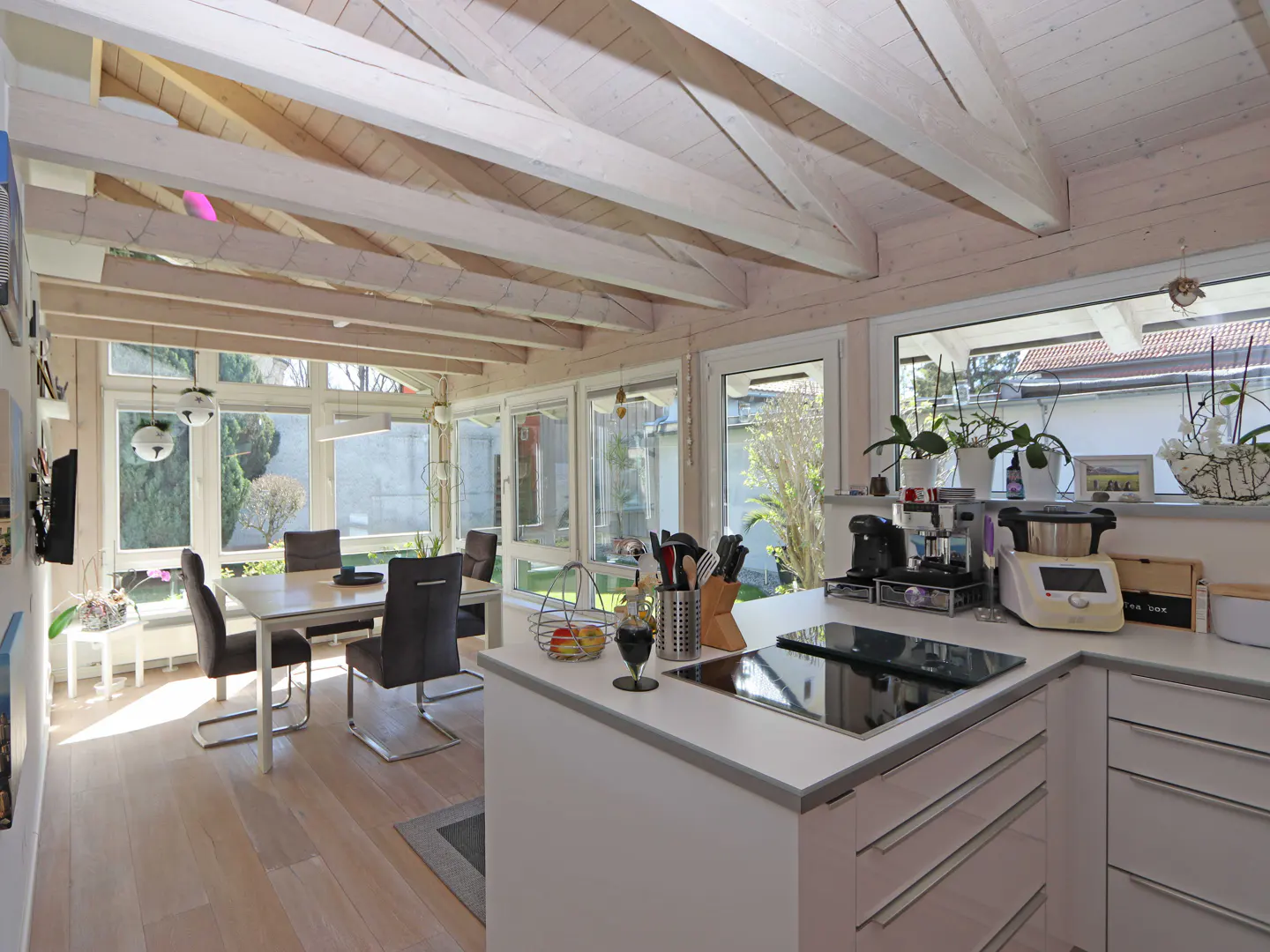 Modern kitchen and dining area with white vaulted ceilings, light wood floors, and large windows overlooking a garden.