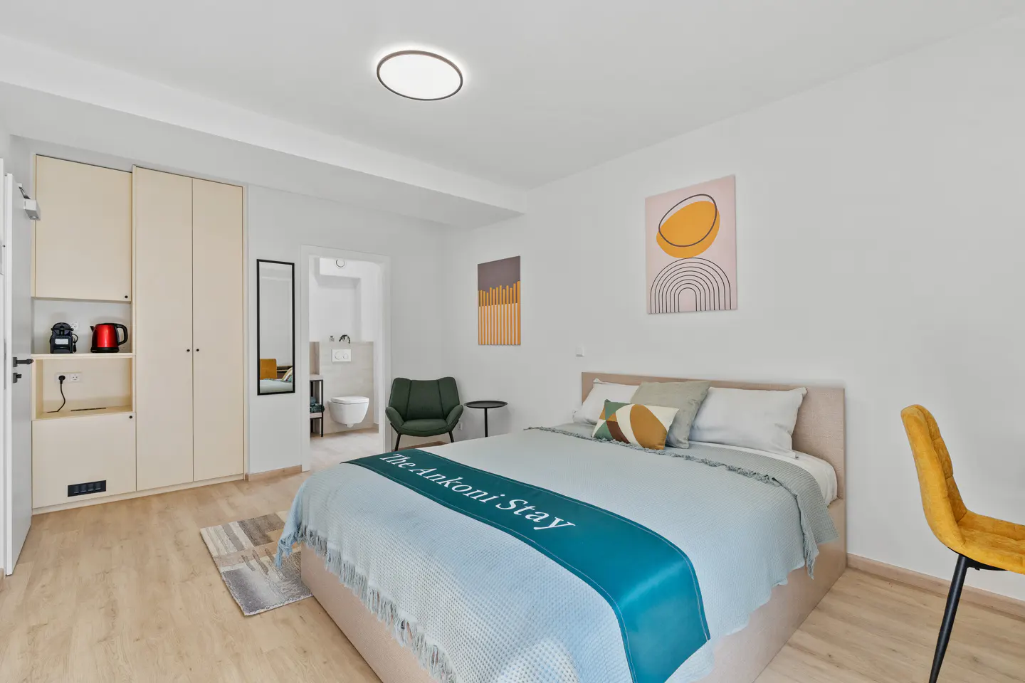 Bright bedroom with a bed, yellow chair, and modern art. A bathroom is visible through an open door.