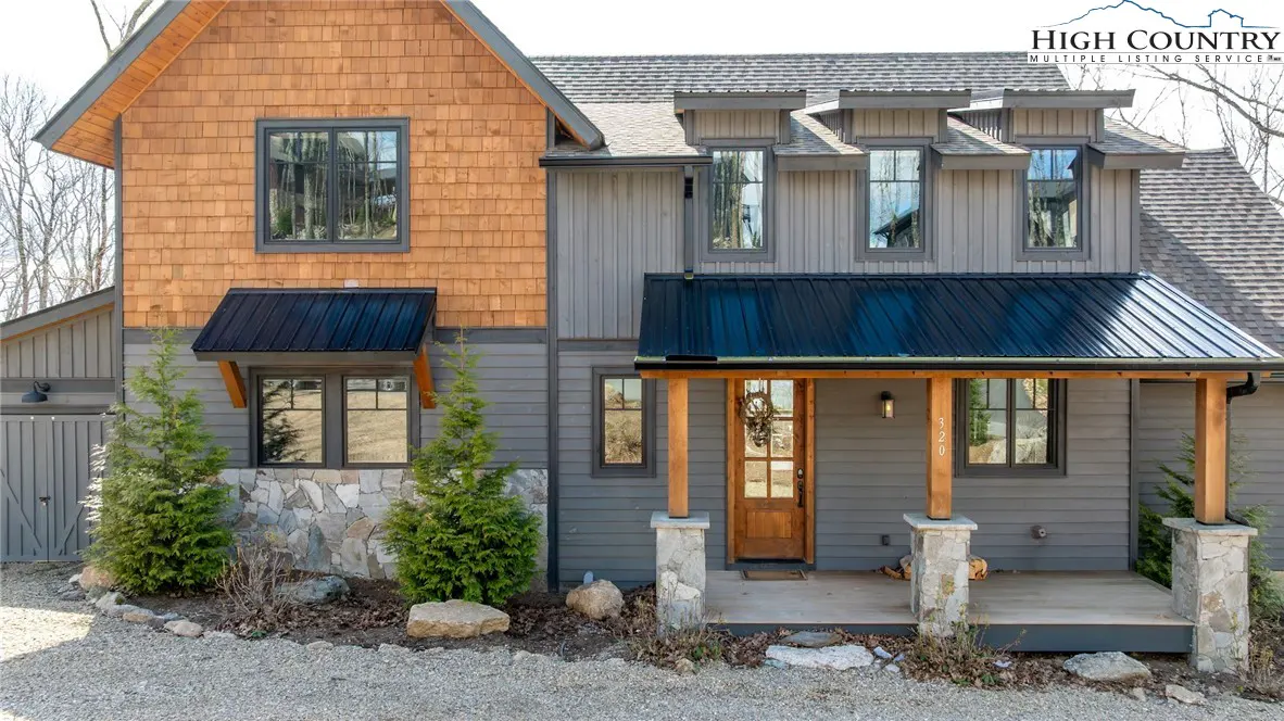 Exterior view of a two-story house with gray siding, wood shingles, black metal roof, and stone accents. A wooden porch covers the front door.