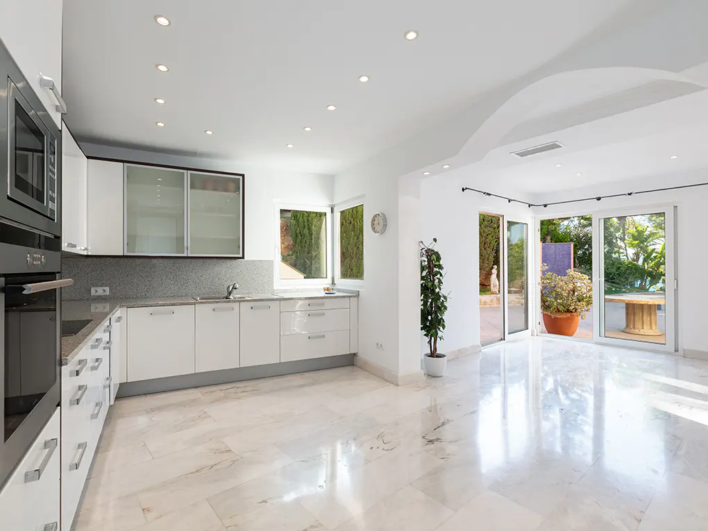 Bright, modern kitchen with white cabinets, marble floors, and stainless steel appliances. Sliding glass doors lead to a patio with lush greenery.