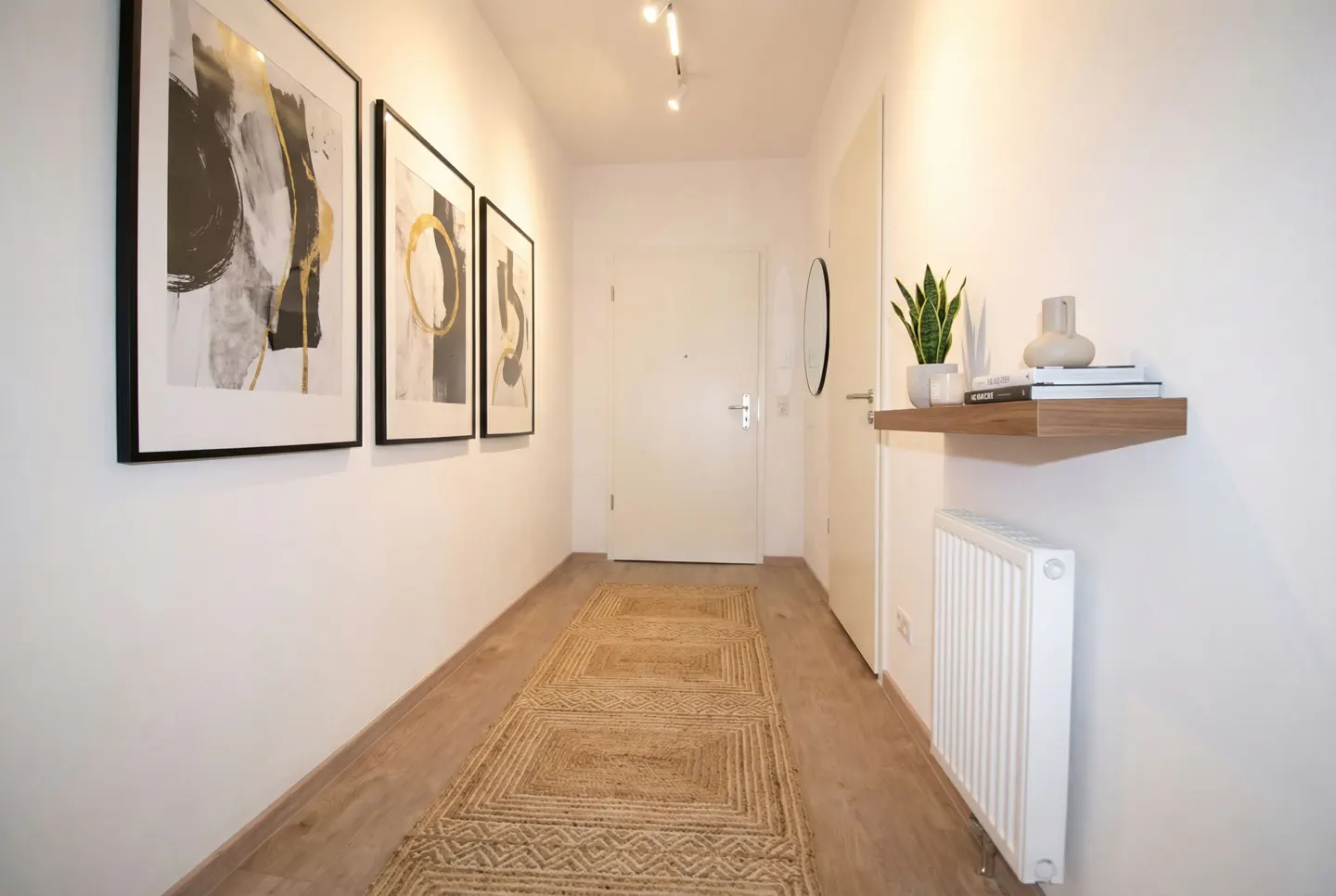 Hallway with white walls, wood floor, and a jute rug. Three framed art pieces hang on the left wall. A floating shelf with decor is on the right.