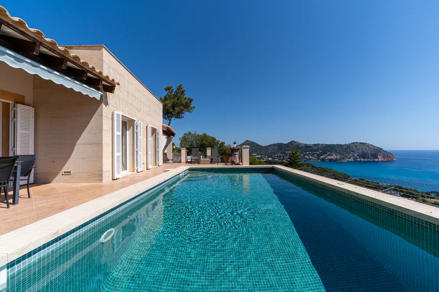 Luxury villa with a turquoise tiled pool overlooking the ocean and mountains under a clear blue sky.