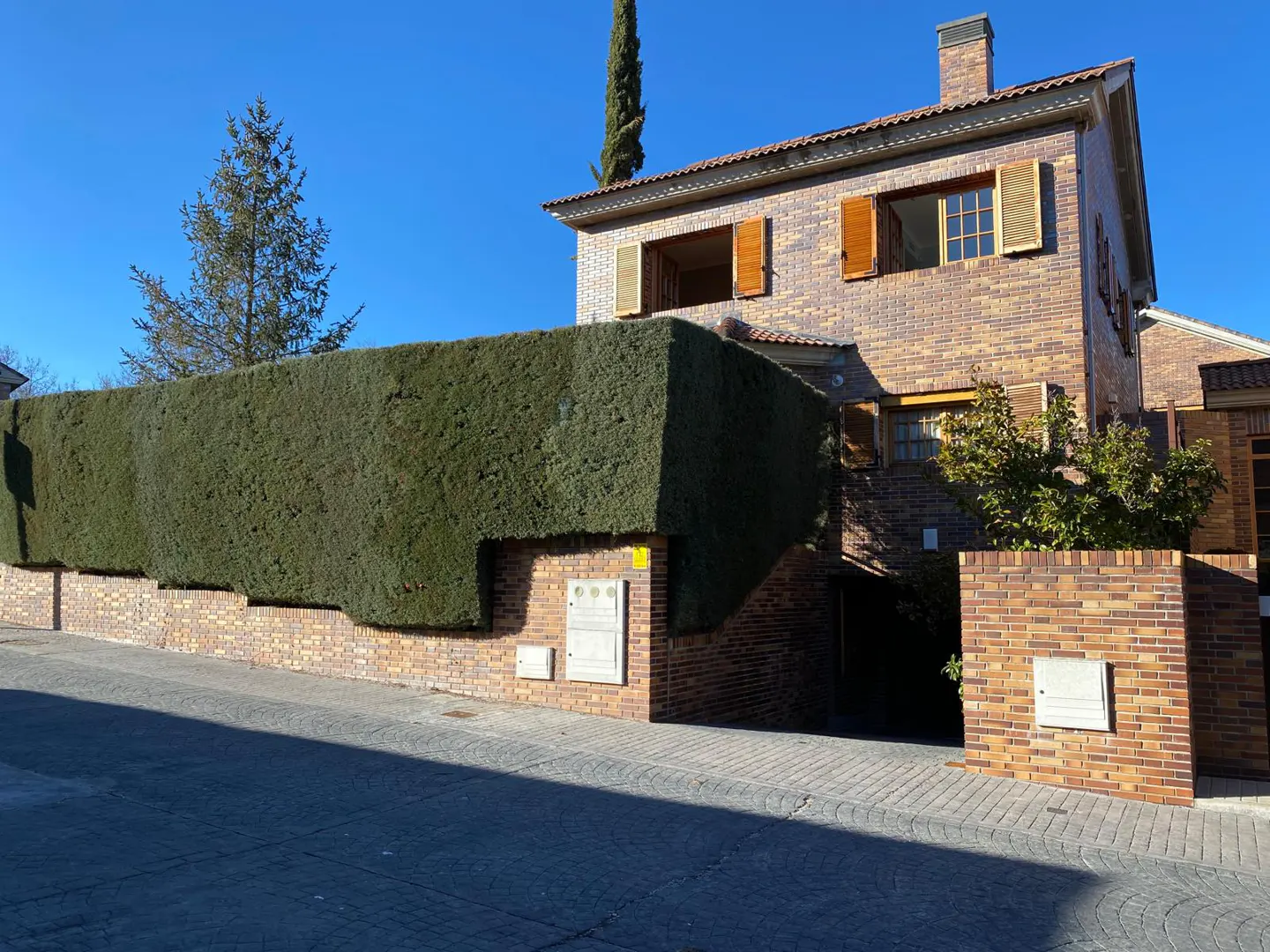 Two-story brick house with brown shutters, a green hedge, and a blue sky.