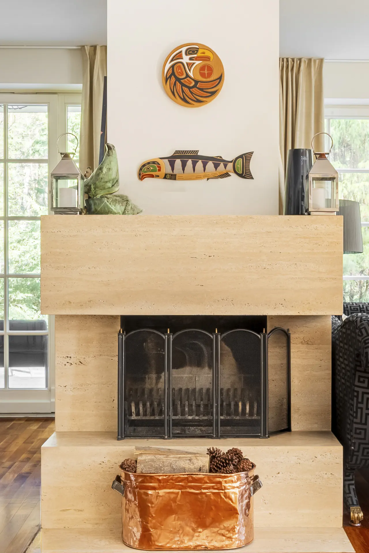 Living room fireplace with tan stone, black metal screen, and copper log holder. Native American art hangs above.