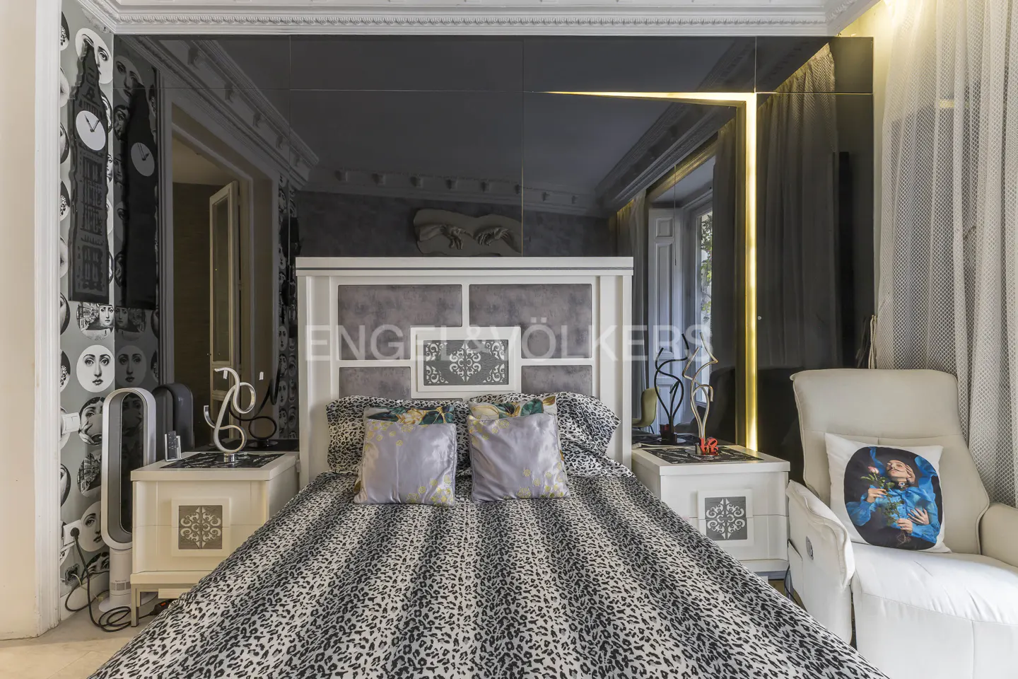 Bedroom with a leopard print bedspread, white headboard, mirrored ceiling, and white armchair with a blue portrait pillow.