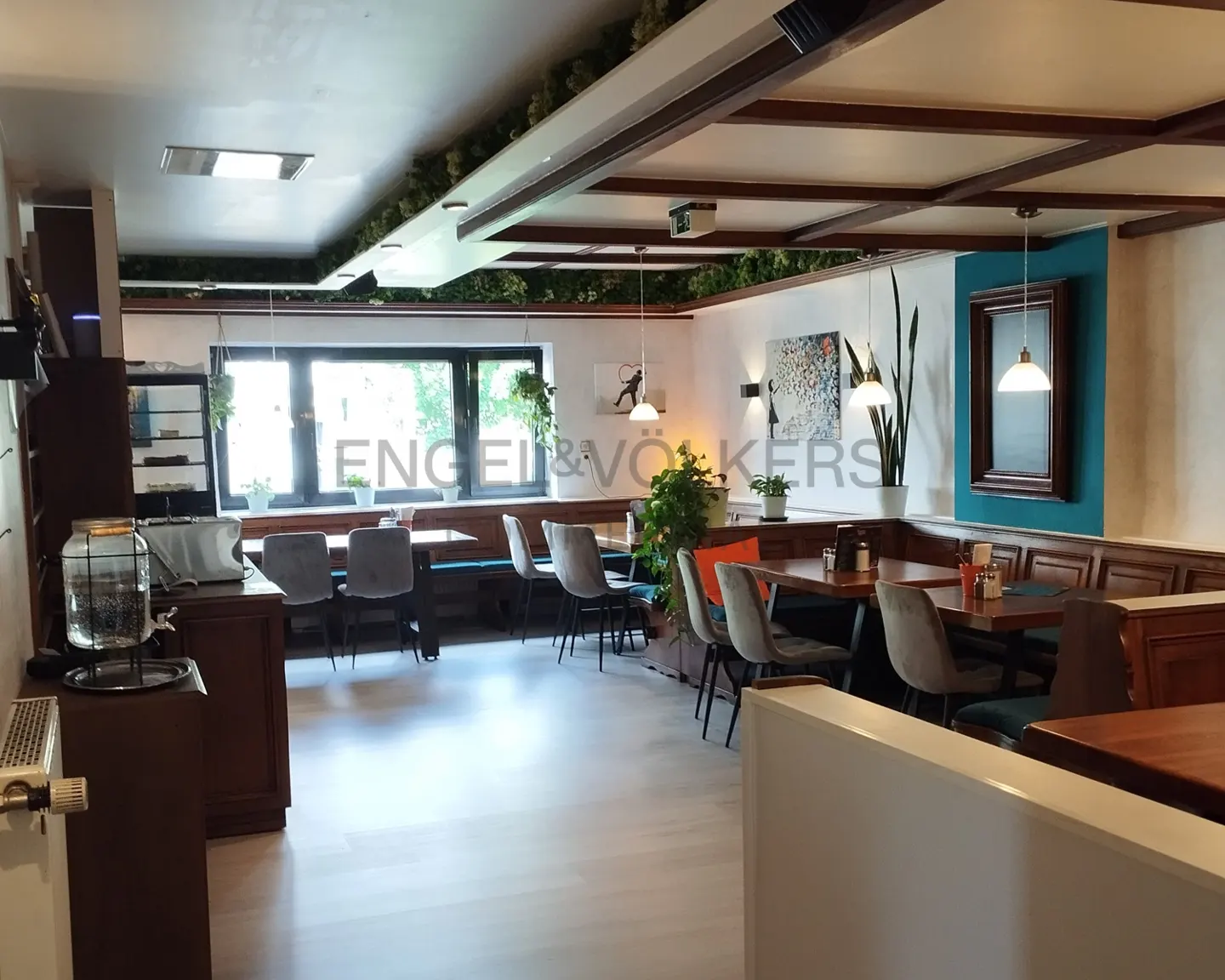 Interior view of a restaurant with tables, chairs, and booth seating. The ceiling has wooden beams and greenery. Engel & Völkers logo is visible.
