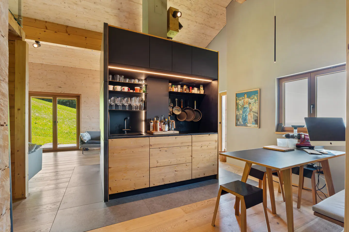 A modern kitchen with black cabinets, wooden drawers, and open shelving. A table with a computer sits nearby. A window overlooks a green field.