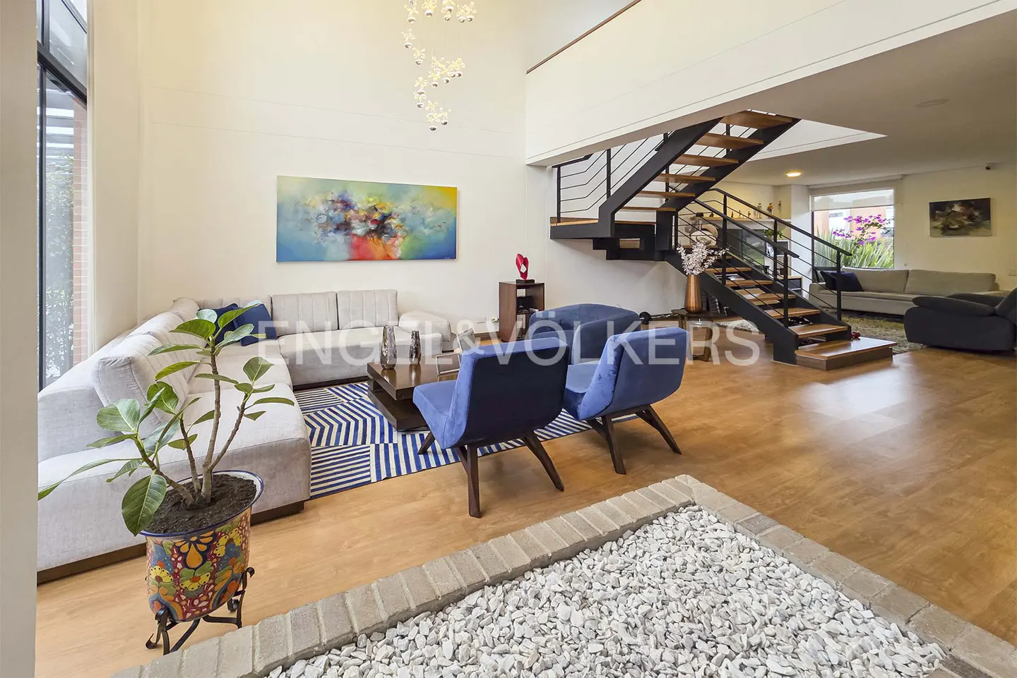 Bright, modern living room with a gray sectional sofa, blue chairs, and a wooden staircase. An abstract painting hangs on the wall.