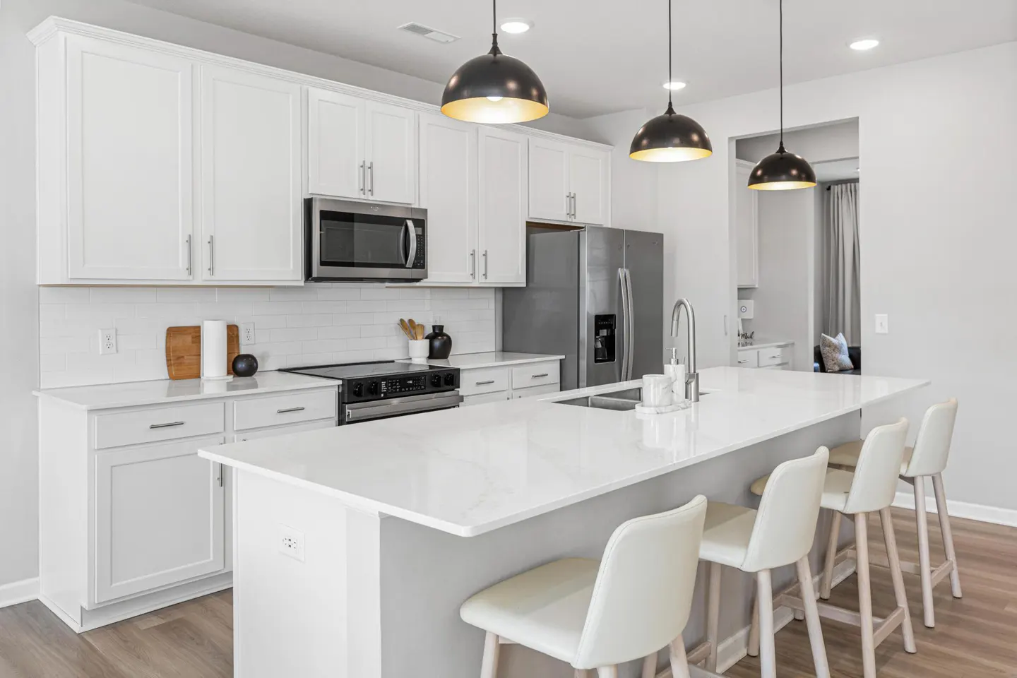 Bright kitchen with white cabinets, stainless steel appliances, and a large island with four white chairs. Black pendant lights hang above the island.