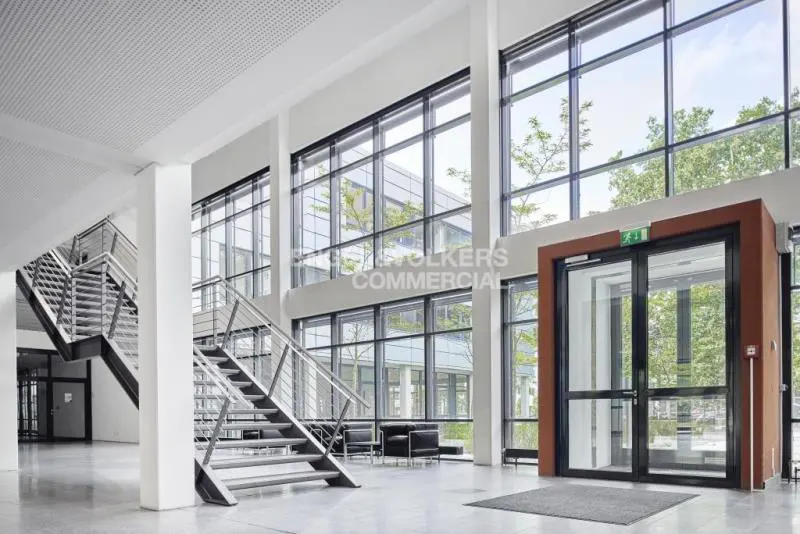 Bright, modern commercial lobby with large windows, stairs, and an exit door. The interior is white and gray, with natural light.