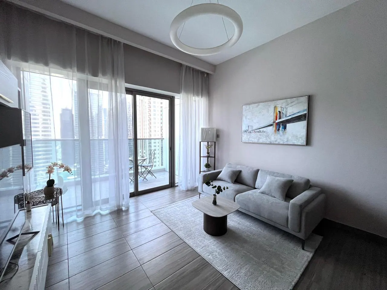 Living room with gray sofa, rug, and balcony with city view. Modern art and circular light fixture. Neutral color palette.