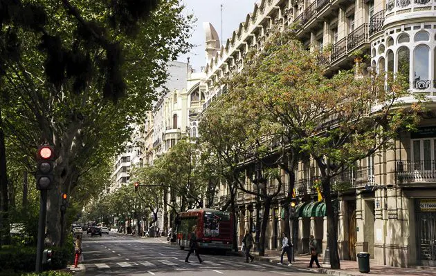 A tree-lined city street with a red bus, pedestrians crossing, and ornate, light-colored historic buildings.