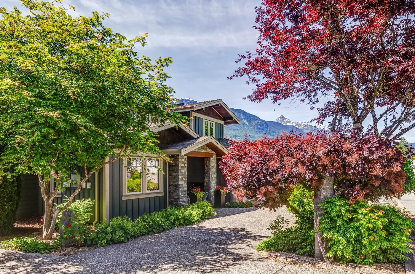 A modern home with dark siding and stone accents, framed by green and red trees with a mountain view in the background.