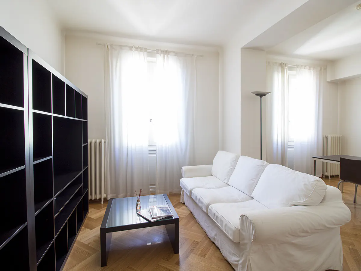 Bright living room with white walls, parquet floor, black bookcase, white sofa, and black coffee table. Windows with sheer white curtains.