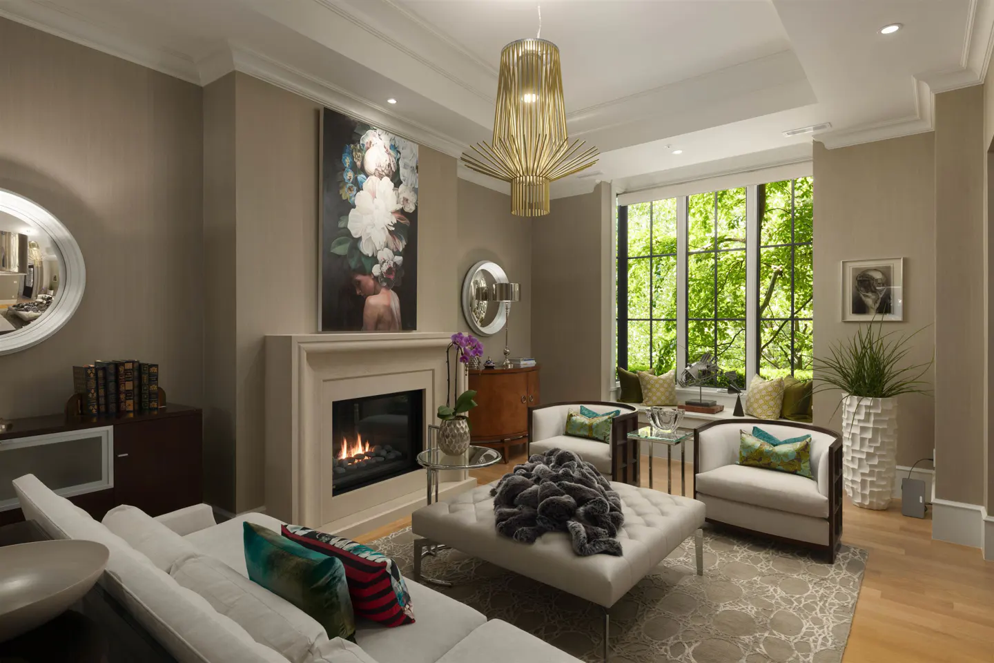 A living room with a fireplace, white sofa, and two chairs. A large window shows green trees. A modern chandelier hangs from the ceiling.