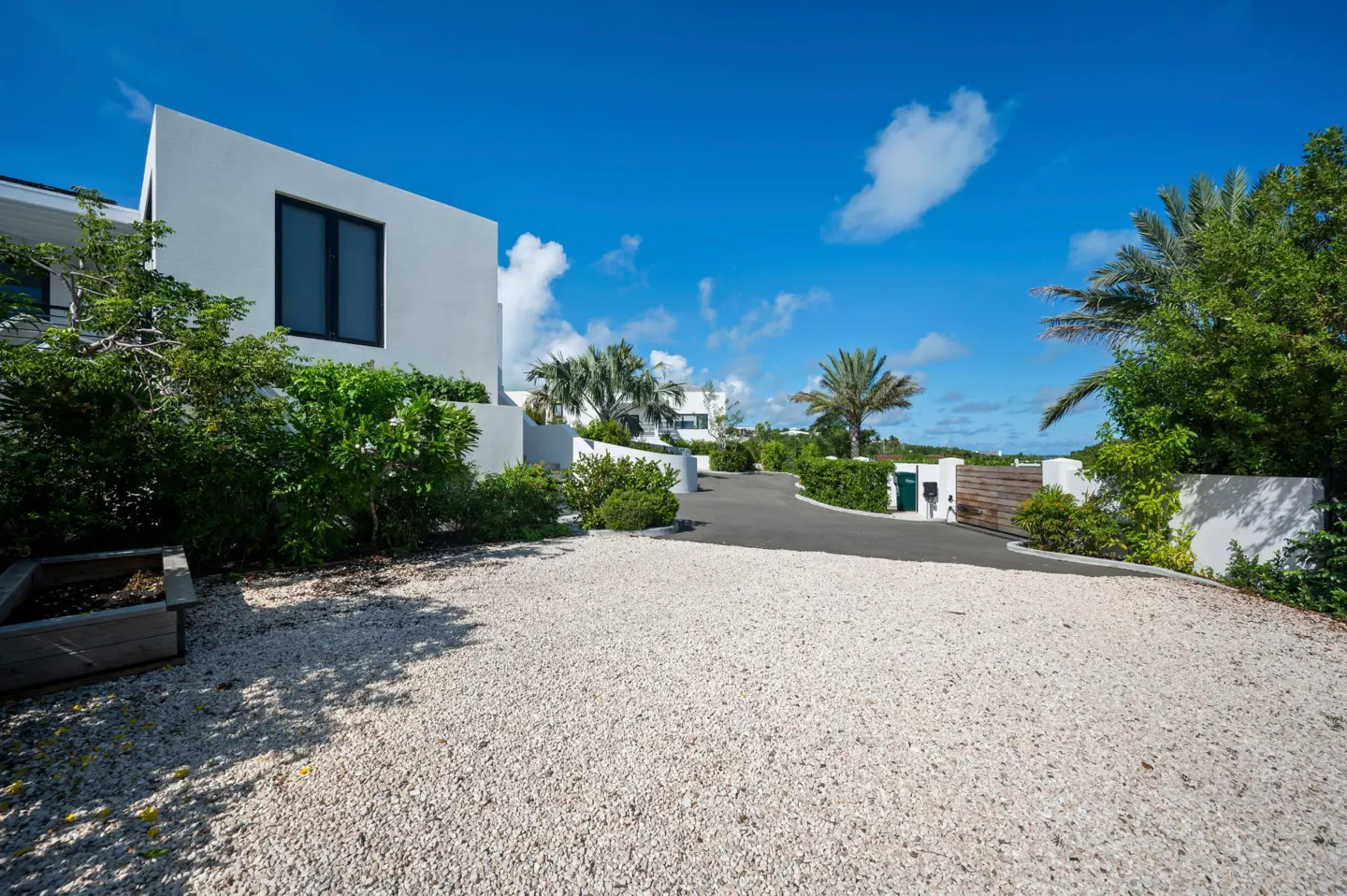Exterior view of a modern white house with a gravel driveway, lush greenery, and a bright blue sky.