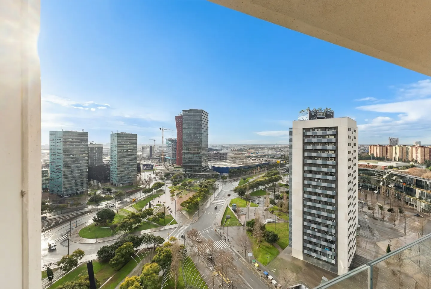 Elevated view from a balcony overlooking a modern city skyline with tall buildings, green parks, and streets under blue sky.