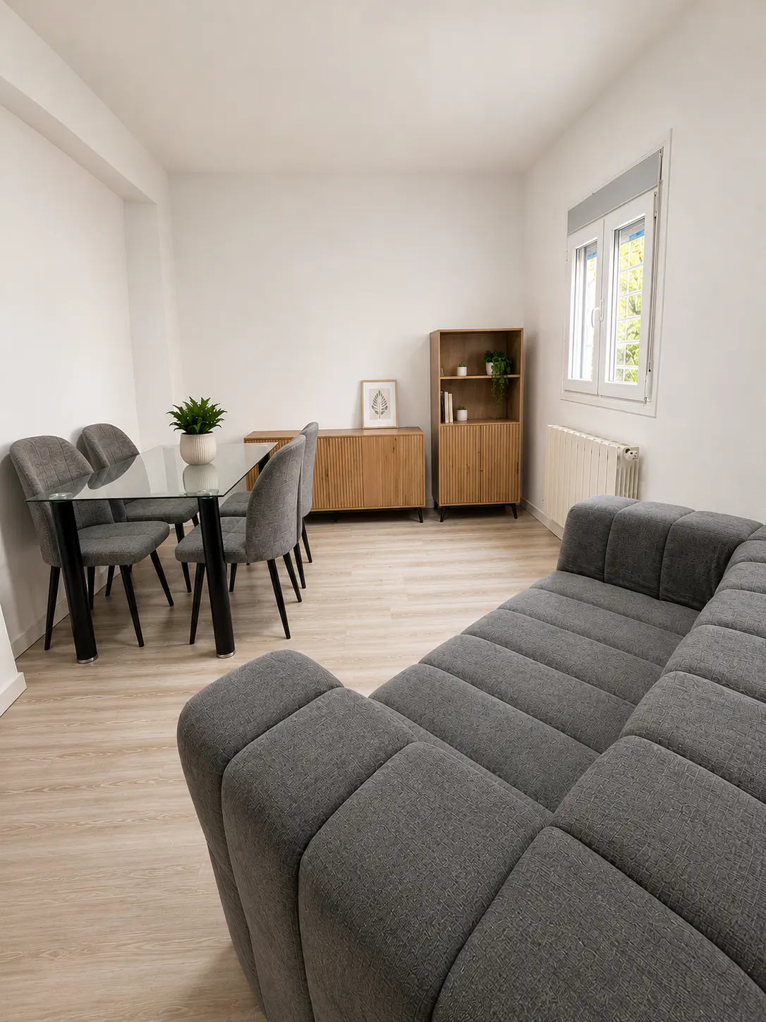 Modern living room with a grey sectional sofa, glass dining table, four grey chairs, and light wood storage furniture.