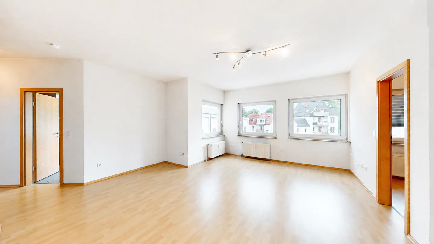 Bright, empty room with wood floors, white walls, and three windows overlooking a neighborhood. A door is open to another room.