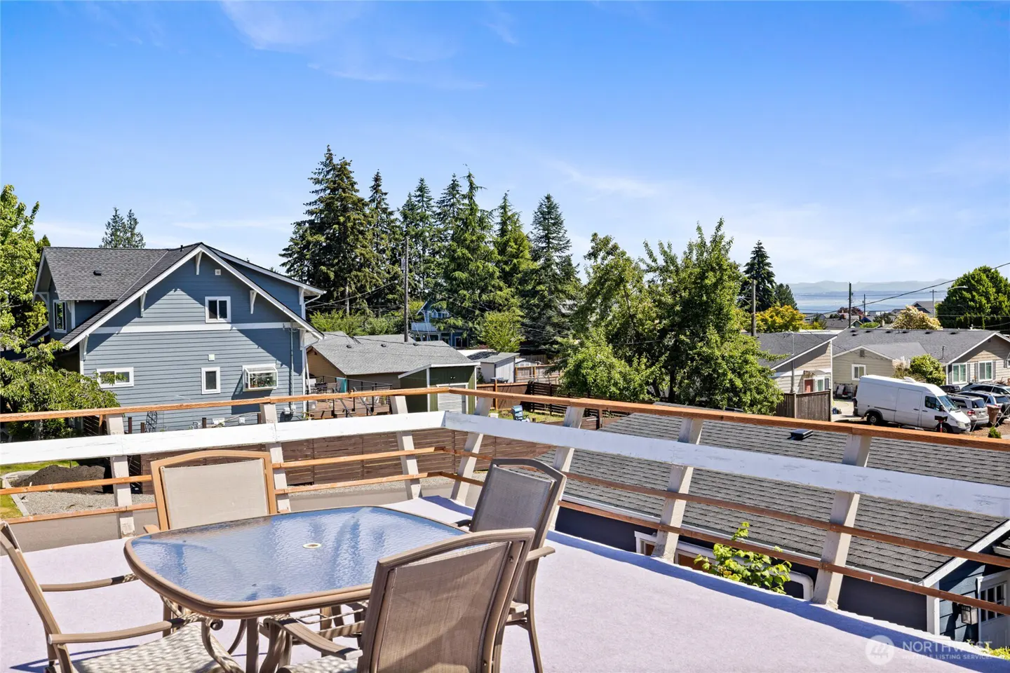 Rooftop deck with a glass-top table and chairs, overlooking a neighborhood with blue houses, trees, and a distant water view.