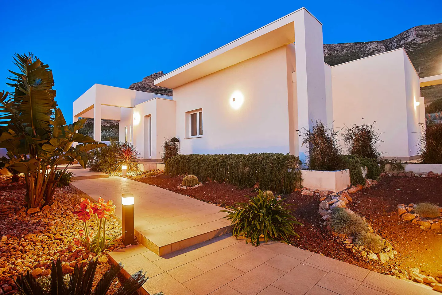 Modern white house at dusk with a stone walkway, garden lights, and lush landscaping. Mountain in the background.