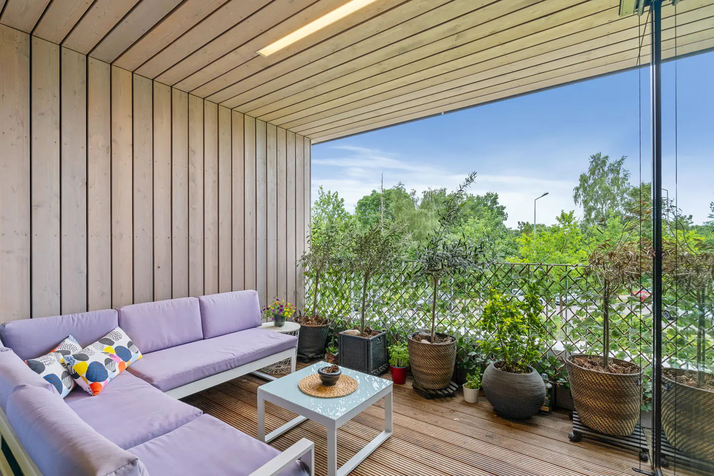 Modern balcony with light wood walls, a lavender sectional sofa, a small white table, and various potted plants.