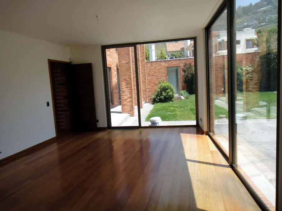 An empty room with hardwood floors and large windows overlooking a brick patio and green lawn.