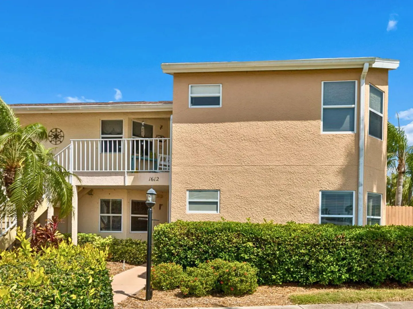 Two-story tan condo building with white balcony and trim, number 1612, green bushes, and blue sky.
